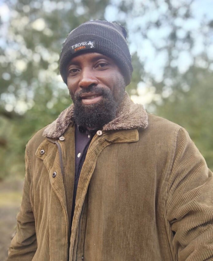 Man outdoors wearing a black knit beanie and brown corduroy jacket with a furry collar, smiling at the camera with trees in the blurred background, chilly weather look.