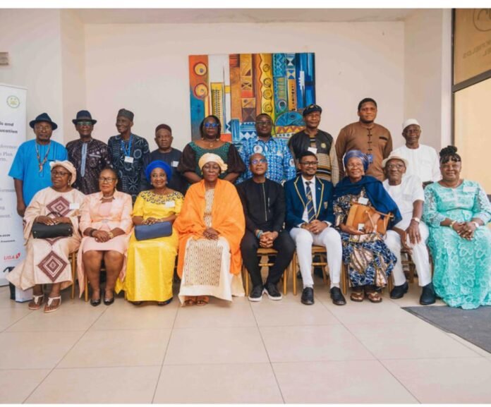 Group of about 18 people posing for a formal group photo indoors, seated in front row and standing behind, with a colorful abstract painting on the wall and a banner to the left; some wear traditional attire and others in suits or dresses.