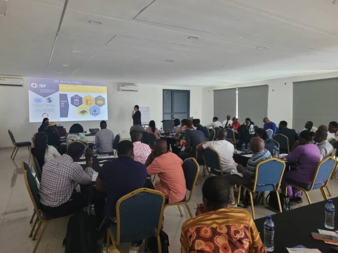 Conference room workshop: presenter at the front addressing a large audience seated around tables with a projector screen overhead.