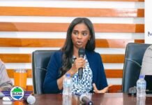 SLCAA DG Reaffirms Safety Priority as Sierra Leone Launches Nationwide NavAIDs Calibration A woman in a blue blazer speaks into a handheld microphone at a panel, seated at a wooden table with water bottles and a logo on the left.