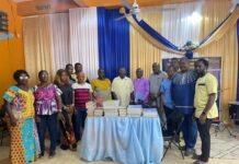 BSSL Distributes Advanced Biblical Texts to Colleges Across Freetown, Bo, Makeni and Kono Group of people in colorful outfits posing behind a table with stacked books at a community event with blue and gold drapes.
