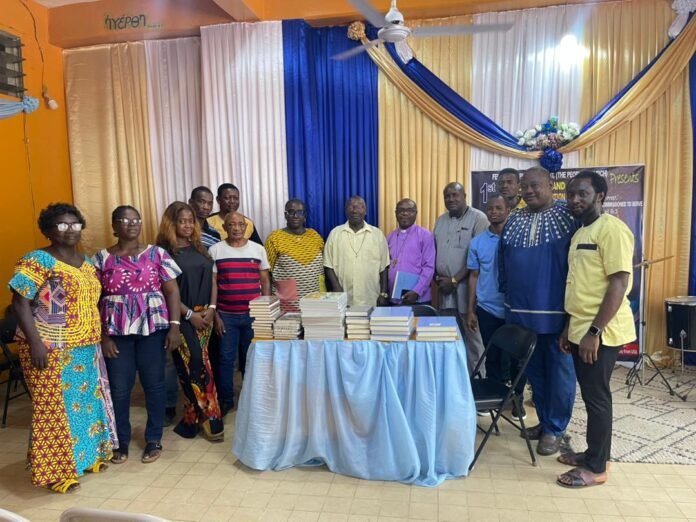 Group of people in colorful outfits posing behind a table with stacked books at a community event with blue and gold drapes.