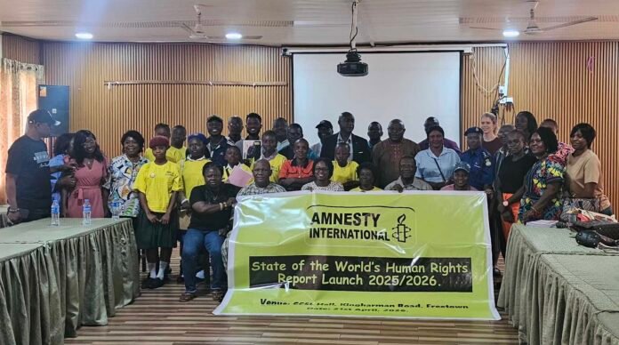 Group of diverse people posing in a room with an Amnesty International banner for the State of the Worldb4s Human Rights Report Launch 2025/2026Reader-friendly version.