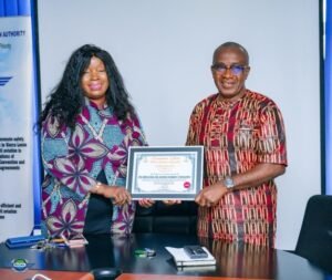 Two colleagues in an office present a framed certificate to each other: the woman on the left wears a purple patterned dress, the man on the right wears a red patterned shirt and glasses, standing by a desk with papers and a calculator.