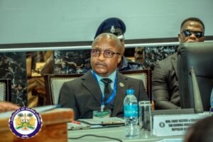 Man in a suit and glasses with a blue lanyard sits at a conference table, water bottle and nameplate in front, Judiciary of Sierra Leone emblem nearby.