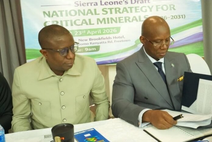 Two officials sign documents at a conference table with a banner for Sierra Leone's National Strategy for Critical Minerals in the background.