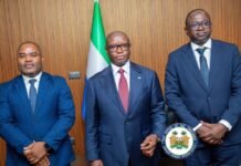 World Bank Executive Directors Commend Sierra Leone’s Reforms, Pledge Continued Partnership Three formally dressed men stand side by side in a government setting, with a flag behind them and an official seal in the foreground.