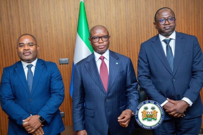 Three formally dressed men stand side by side in a government setting, with a flag behind them and an official seal in the foreground.