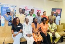 TOES-SL Hosts Youth Roundtable Discussion on Empowering Sierra Leone Youths for National Development Group of 12 people posing for a photo at a conference, seated in front row with banners in the background and an American flag banner on the right.