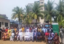 ECOSOC Convenes Multi-Stakeholder Dialogue on Democratic Governance in Sierra Leone Large group of people posing for a photo outdoors, seated and standing in front of palm trees and a building, suggesting a community or conference event.