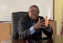 Health Alert, WASHNet Commend Government Over GAVI Co-Financing Approval Man in a dark suit and glasses sits at a desk, speaking with animated hand gestures during a meeting.