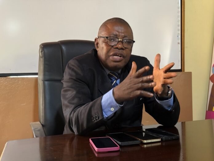 Man in a dark suit and glasses sits at a desk, speaking with animated hand gestures during a meeting.