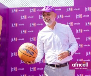 Smiling man in a white shirt and purple cap holds an orange basketball in front of a purple Africell event backdrop.