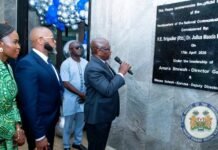 President Bio Commissions State-of-the-Art NatCA Headquarters, Reinforces Digital Transformation Agenda Dignitaries at a plaque unveiling ceremony; a man in a suit speaks into a microphone as others in formal attire and a woman in green watch, with balloon decorations in the background.
