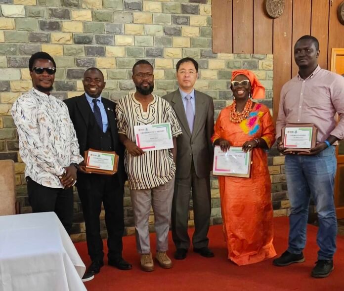 Six adults posing indoors, each holding a certificate; a woman in bright orange traditional dress with headwrap stands among men in formal or traditional attire.