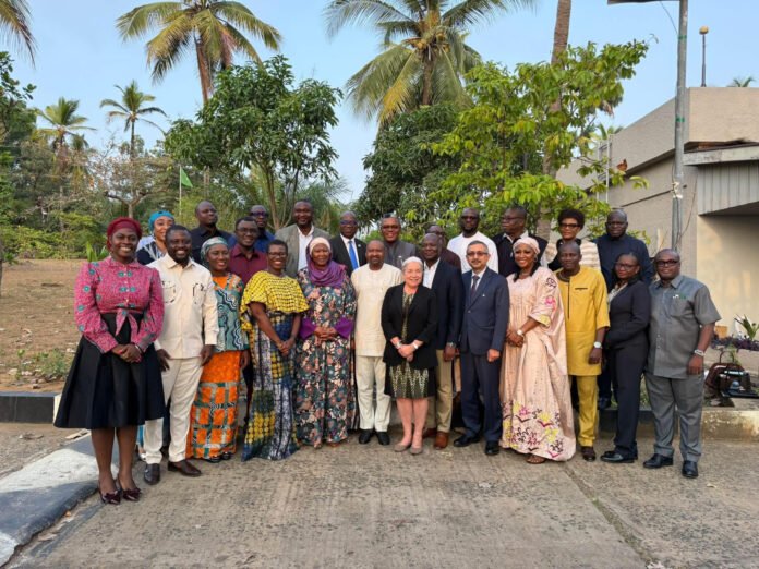 Group of diverse men and women in colorful traditional and formal clothing posing for a group photo outdoors among palm trees and a building backdrop, on a sunny day.