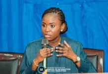 Sierratel Set for Comeback as Government Adopts MVNO Model with Africell Woman in teal blouse speaks into a handheld microphone at a panel, with a blue curtain backdrop and a Sierra Leone government ministry logo on the podium.