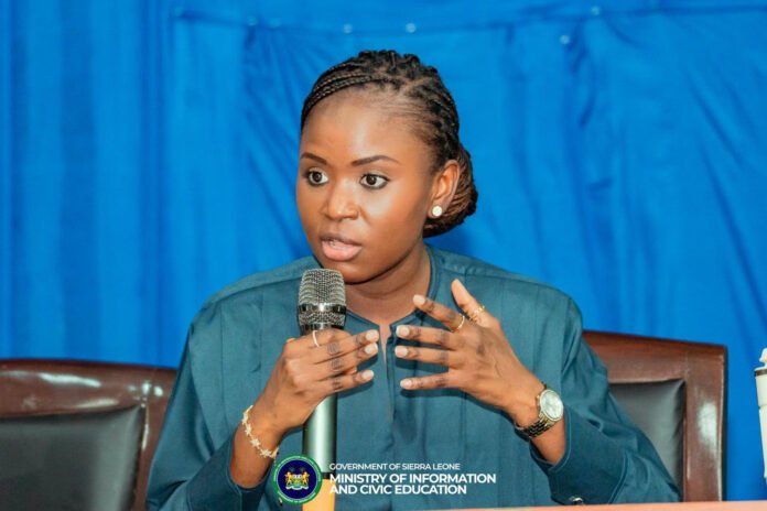 Woman in teal blouse speaks into a handheld microphone at a panel, with a blue curtain backdrop and a Sierra Leone government ministry logo on the podium.