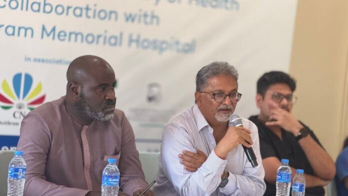 Panel of three men at a conference table; center man speaks into a microphone while others listen, with water bottles on the table and a blurred banner in the background.