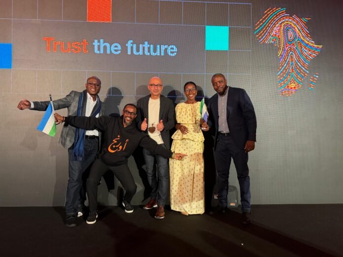 Six diverse adults posing on a stage in front of a backdrop reading 'Trust the future' with a colorful fingerprint graphic, some holding small flags and smiling.