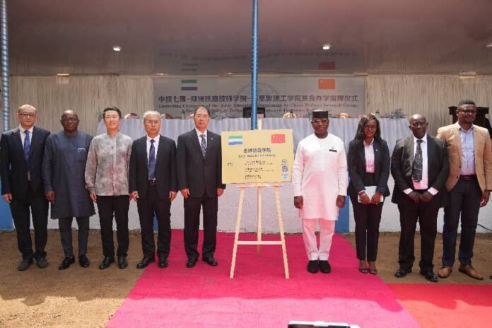 Group of formally dressed people standing on a pink carpet during an unveiling ceremony, with a signboard on an easel at the center and a banner in the background.