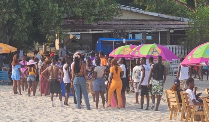 Crowd of people in swimsuits standing and chatting on a sunny beach near colorful umbrellas and outdoor seating.