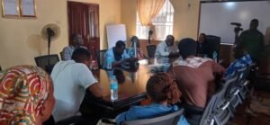 Group of people seated around a conference table in a meeting room, with cameras and a projector present, discussing.