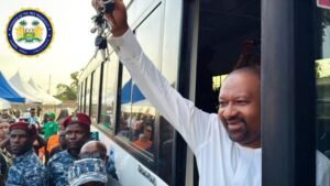 Man in a white traditional outfit waves from an open bus door as a crowd and security personnel look on; official seal appears in the top-left corner.