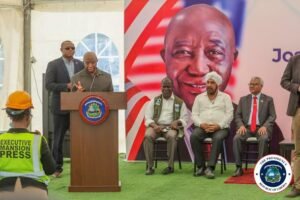Man speaks at a podium with the Liberian presidential seal as officials sit nearby, with a photographer in an Executive Mansion Press vest observing and a large portrait backdrop behind them.