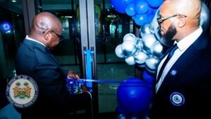 Two men in suits cut a blue ribbon at a glass door entrance during a ceremony, with blue balloons in the background.