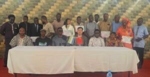 Group of men and women in formal and traditional attire posing with certificates at a ceremony, seated at a long table with two small flags in front.