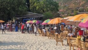 Crowded beach scene with wooden tables under colorful umbrellas and people dining on the sand by trees and a building in the background.