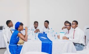Group of students in formal attire seated around a long table with water bottles and documents, engaged in discussion in a bright room with white walls and a blue table runner.