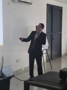 Man in a dark suit and glasses giving a presentation with a microphone in a conference room