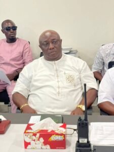 Man in a white embroidered tunic sitting at a desk during a meeting, with tissue box and radio in front of him.