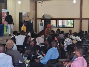 Crowded indoor hall with a stage on the left and a speaker in a yellow shirt, banners nearby, as attendees sit and converse.