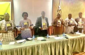 Six people stand behind a long table in a conference room, each holding a copy of a dark blue book with a globe graphic on the cover. They pose for a book launch or presentation event.