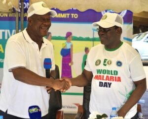 Two men wearing white caps shake hands in a World Malaria Day event, smiling at a booth with a banner behind them.