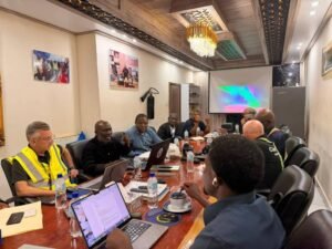 A diverse group of people seated around a long conference table with laptops, water bottles, and coffee cups in a meeting room with a projector screen in the background.
