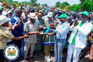 Crowd at a political rally watches as an official hands a ceremonial item to another man, with the presidential seal visible in the corner.