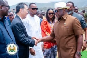 Dignitaries outdoors greet with a handshake; a man in a brown shirt and hat shakes hands with a suited official, while others look on with mountains in the background.