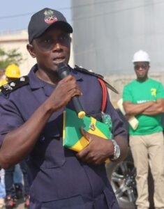 Male police officer in a dark uniform speaks into a handheld microphone, holding a green and yellow folder, with construction workers and a person wearing a white hard hat in the background.