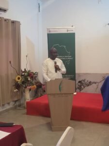 Man in white shirt speaking at a podium with a microphone in a conference room; banner and floral arrangement visible in the background.