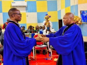 Two clergymen in blue liturgical robes shake hands during a formal ceremony in a colorfully tiled church, with a red carpet, seated audience, and a photographer capturing the moment in the background.
