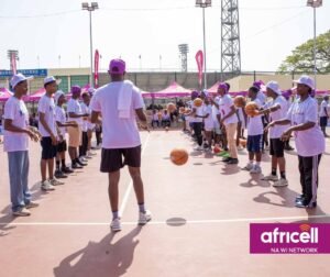 Young players in white shirts and purple caps line up on a basketball court, bouncing balls as part of a community event, with an Africell banner in the corner.
