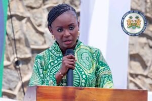 Woman in a green patterned dress speaks into a handheld microphone from a wooden podium at an outdoor formal event, with a government seal in the background.