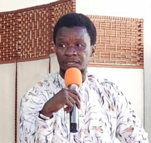 Man in a light patterned traditional outfit holds an orange microphone and speaks indoors.