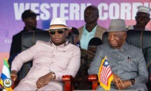 Two men sit on stage in chairs during a formal event; one in a pale pink shirt, white hat and sunglasses, the other in a gray suit with a fedora, with Botswana and U.S. flags in front.