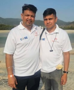 Two men standing together on a sandy beach, wearing white polo shirts with logos and shoulder straps visible in a sunny outdoor setting.