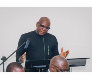 Bald man in black traditional attire speaks at a lectern with a microphone, gesturing with his hands as he addresses an audience.